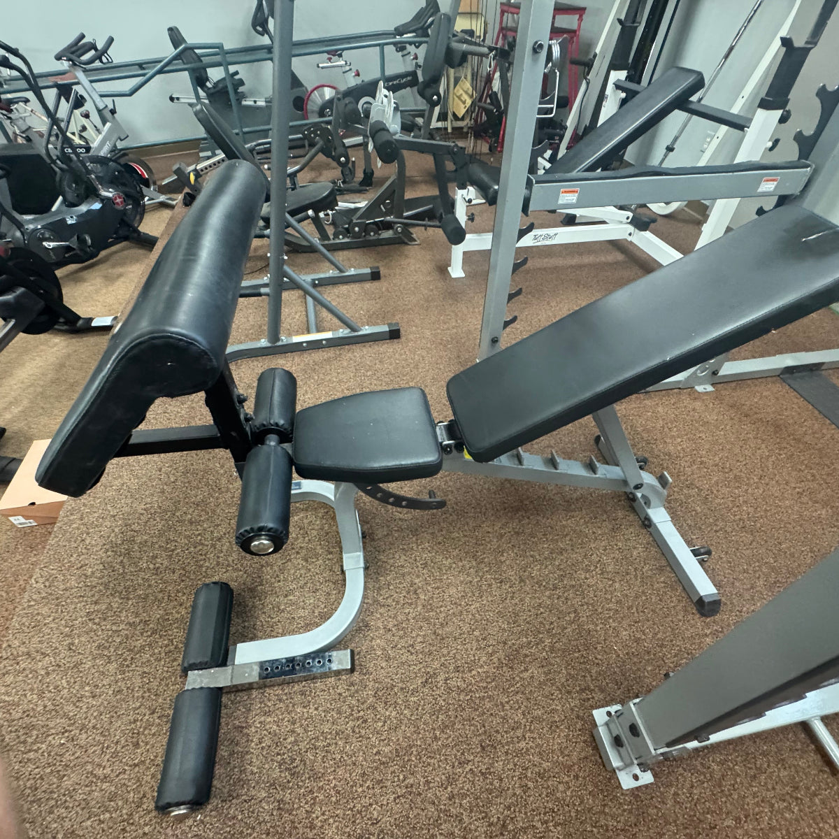 A black and silver adjustable weight bench with a footrest, photographed in a gym setting with other exercise equipment visible in the background.