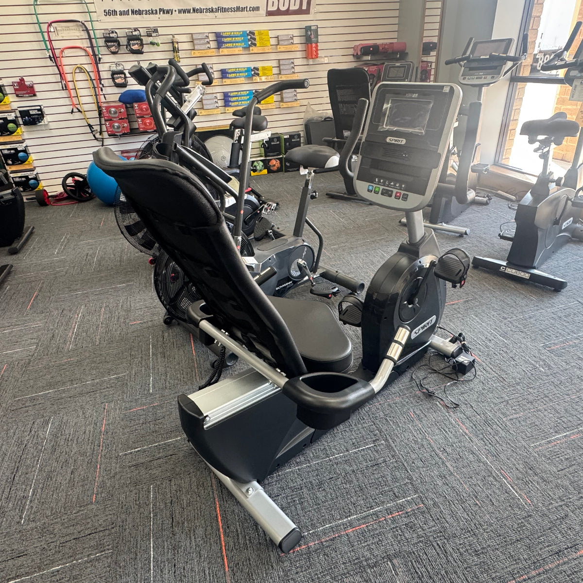 A home gym interior showing a large recumbent exercise bike in the foreground, with other exercise equipment, treadmills, and a wall rack visible in the background.