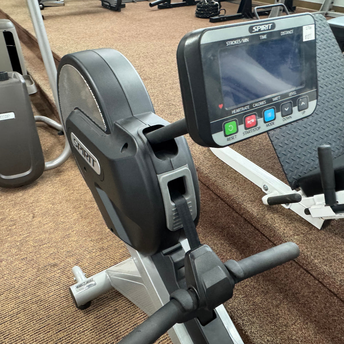 A close-up of a rowing machine in a gym setting, featuring a black and gray flywheel with a digital display screen on the console, set on a brown carpeted floor.
