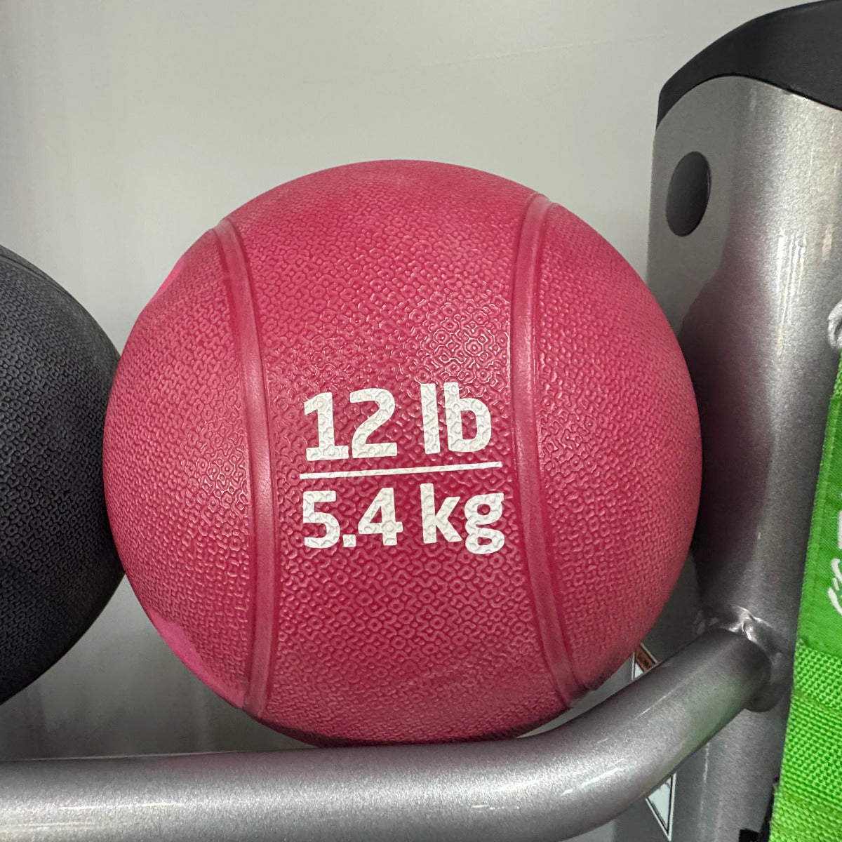 Red medicine ball labeled '12 lb 5.4 kg' on a rack with gym equipment.