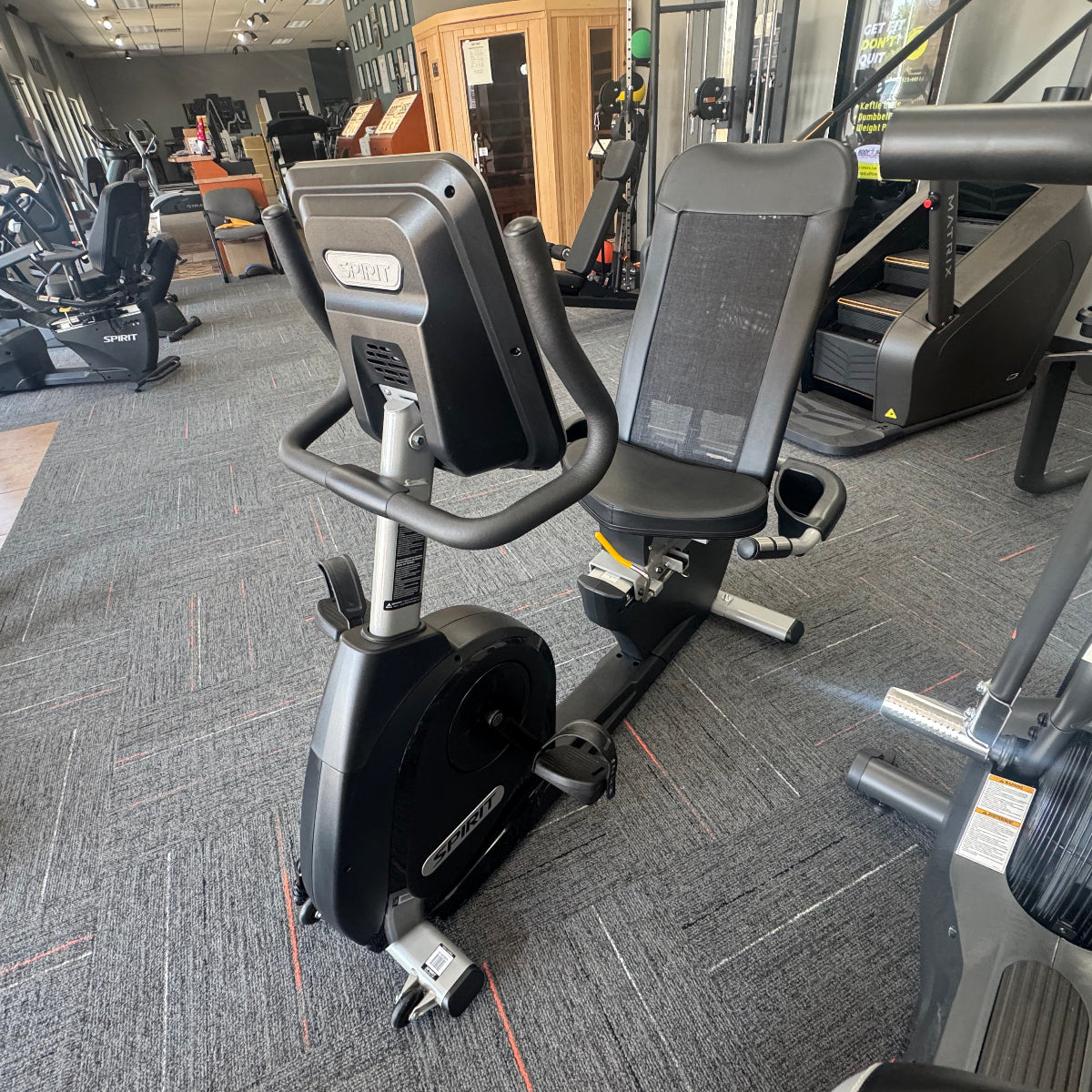 A black and gray indoor stationary bike with an integrated digital display screen, photographed inside a gym with other exercise equipment and a carpeted floor.