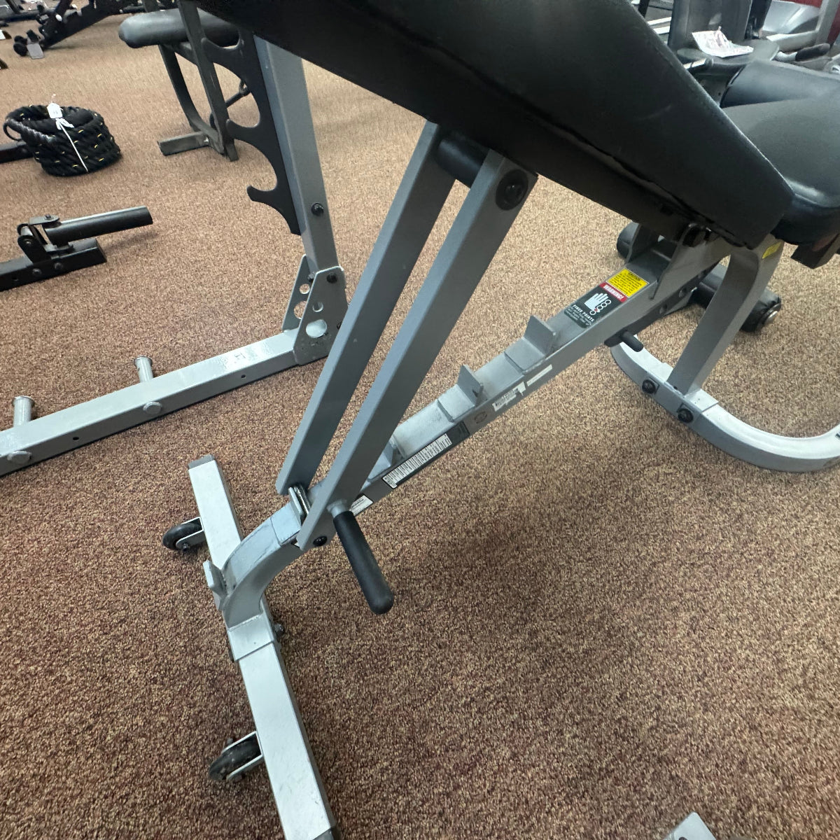 A silver and black adjustable weight bench with a curved backrest, photographed in a gym setting on brown carpet with other exercise equipment visible in the background.