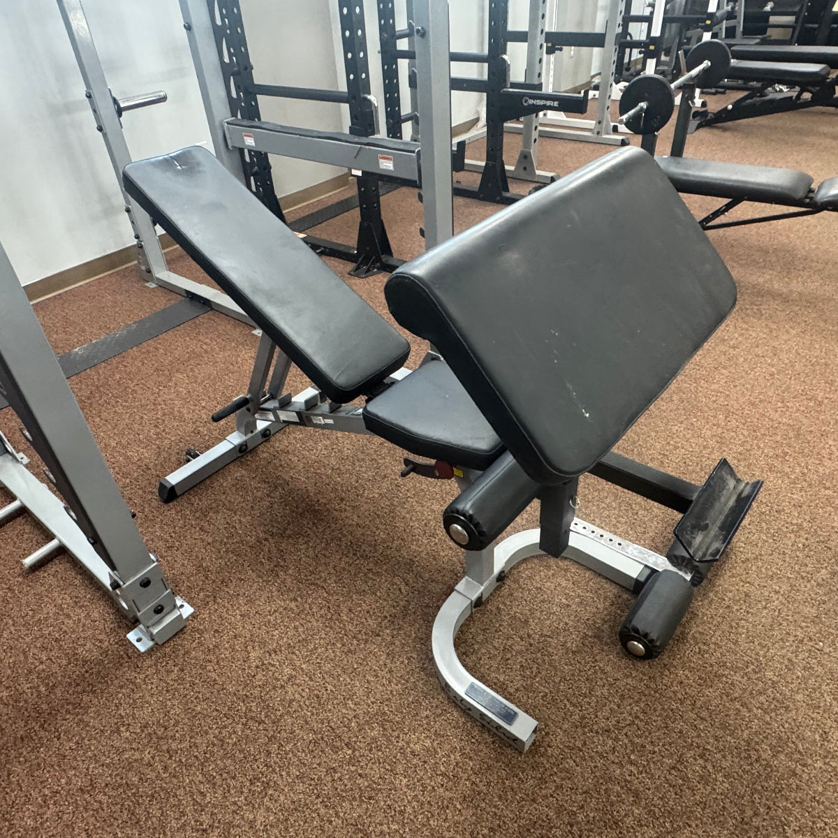 A black adjustable weight bench with a silver metal frame, situated on a brown carpeted gym floor with other exercise equipment in the background.