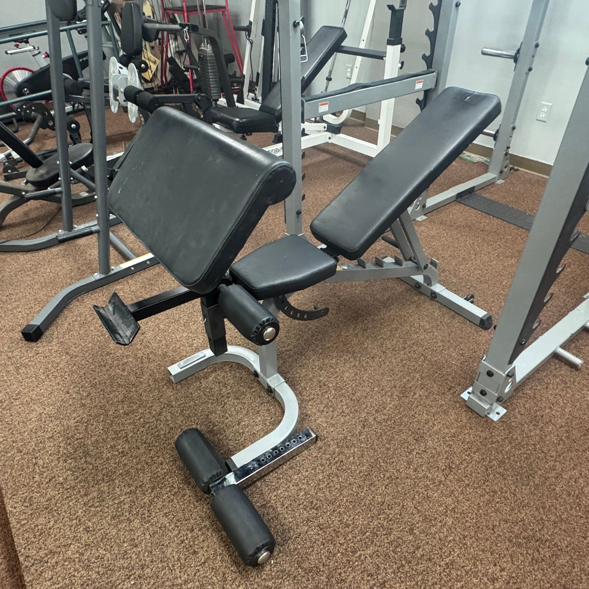 A black and silver adjustable weight bench with a leg curl attachment, photographed in a gym setting with other exercise equipment and brown carpet.