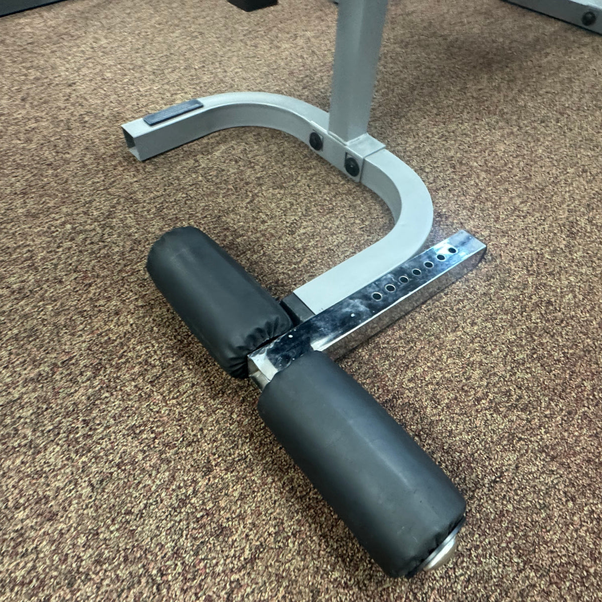 A close-up of a silver and black adjustable weight bench leg support pad on a brown carpeted gym floor.