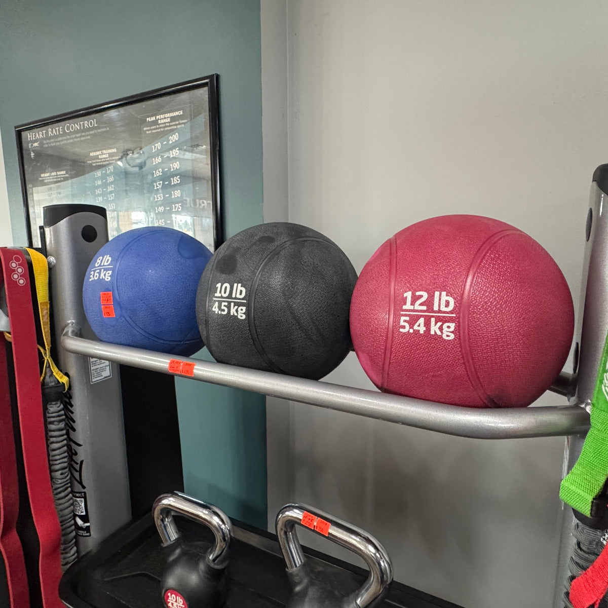 Three exercise balls of different colors and weights on a rack in a gym setting.
