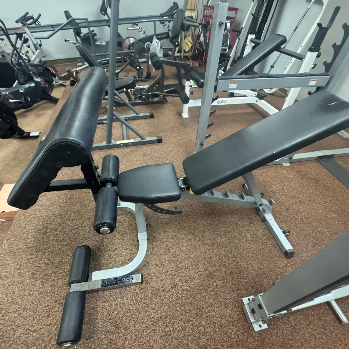 A black adjustable weight bench with a silver metal frame, photographed inside a gym with other exercise equipment and tan carpet visible in the background.