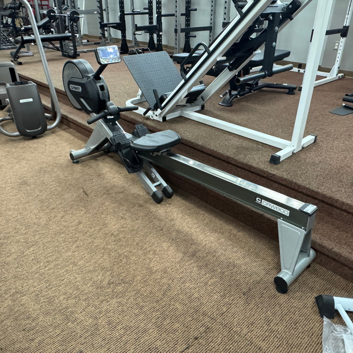 A rowing machine in a gym setting with brown carpet, other exercise equipment, and a white step platform in the background.